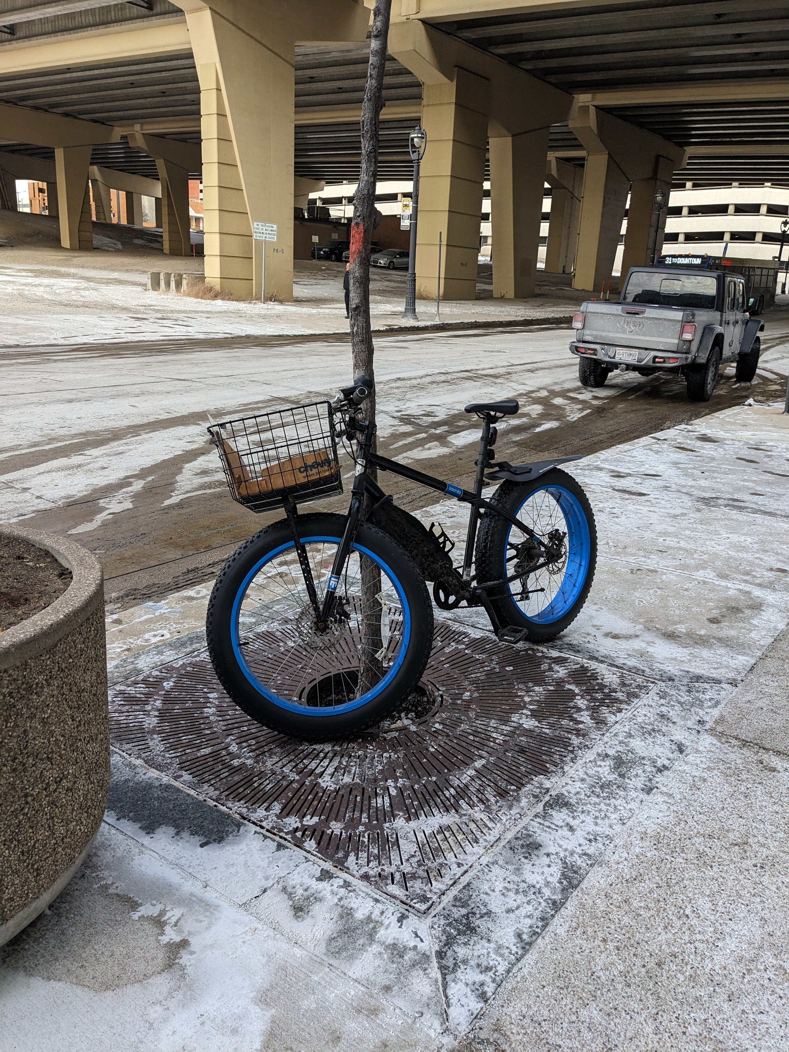 A black fatbike with blue rims and a basket is locked to a small tree on a snow covered sidewalk. A highway underpass is in the background.