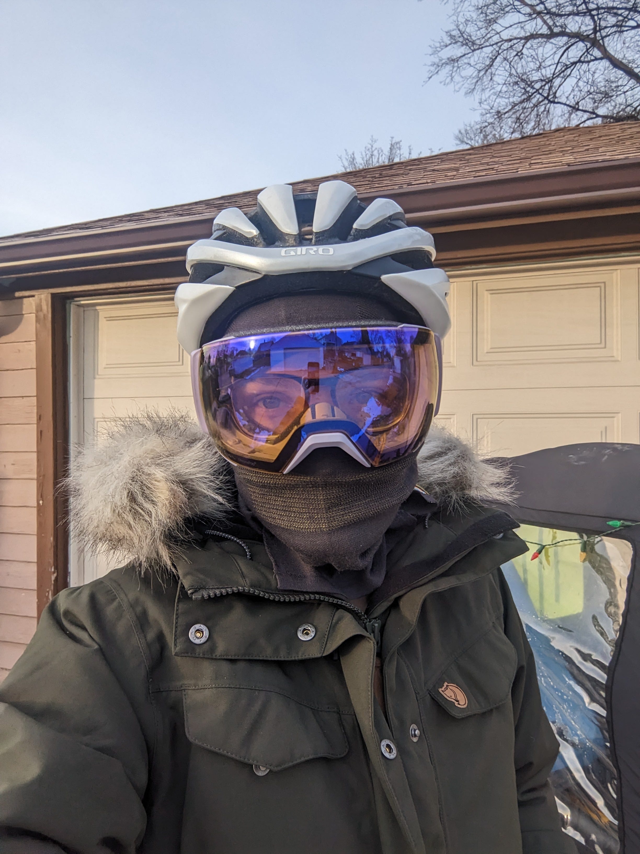 A picture of Anna standing in front of a garage and cargo bike, wearing a heavy winter coat, balaclava, ski goggles, and helmet.