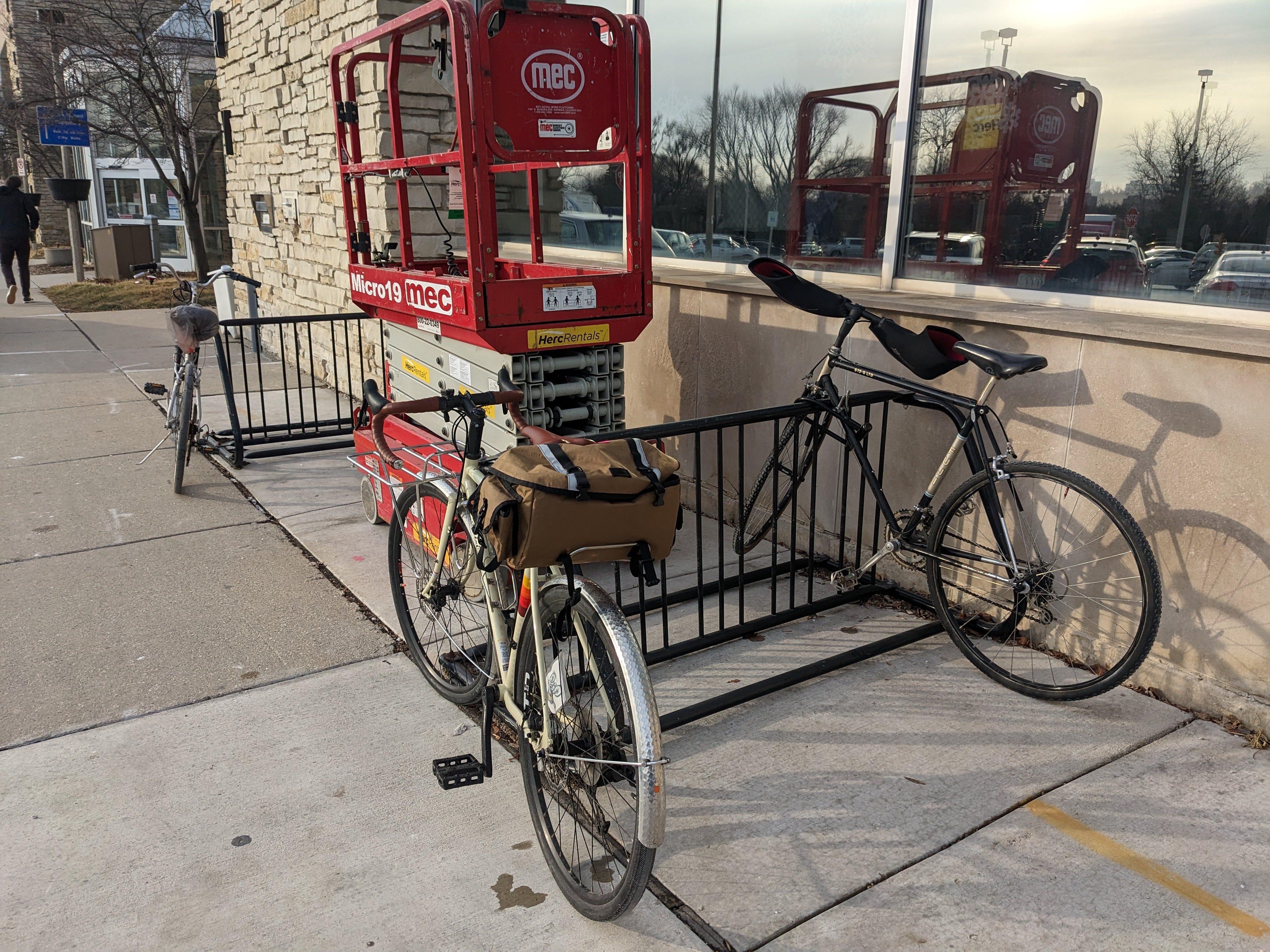 Two "schoolyard" style bike racks outside a lannon stone building with large windows with 3 bikes attached. Between them, a large lift is parked. 